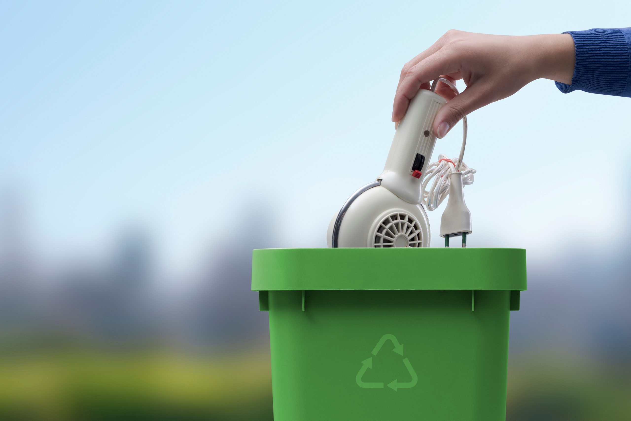 En hand lägger en vit elprodukt i en grön behållare. Grönt landskap i bakgrunden, blå himmel.